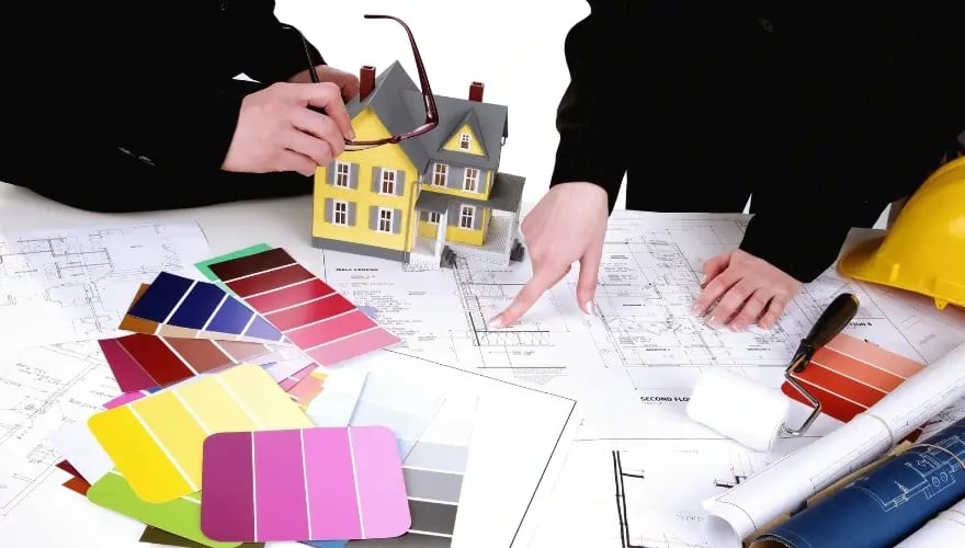 Architekten besprechen Umbaupläne mit Farbmusterkarten und Modellhaus für barrierefreies Bauen.