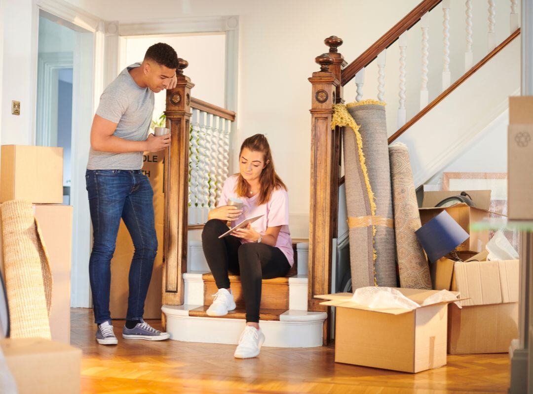 Ein Paar sitzt auf der Treppe eines neuen Hauses mit Umzugskartons und Teppichen im Hintergrund.